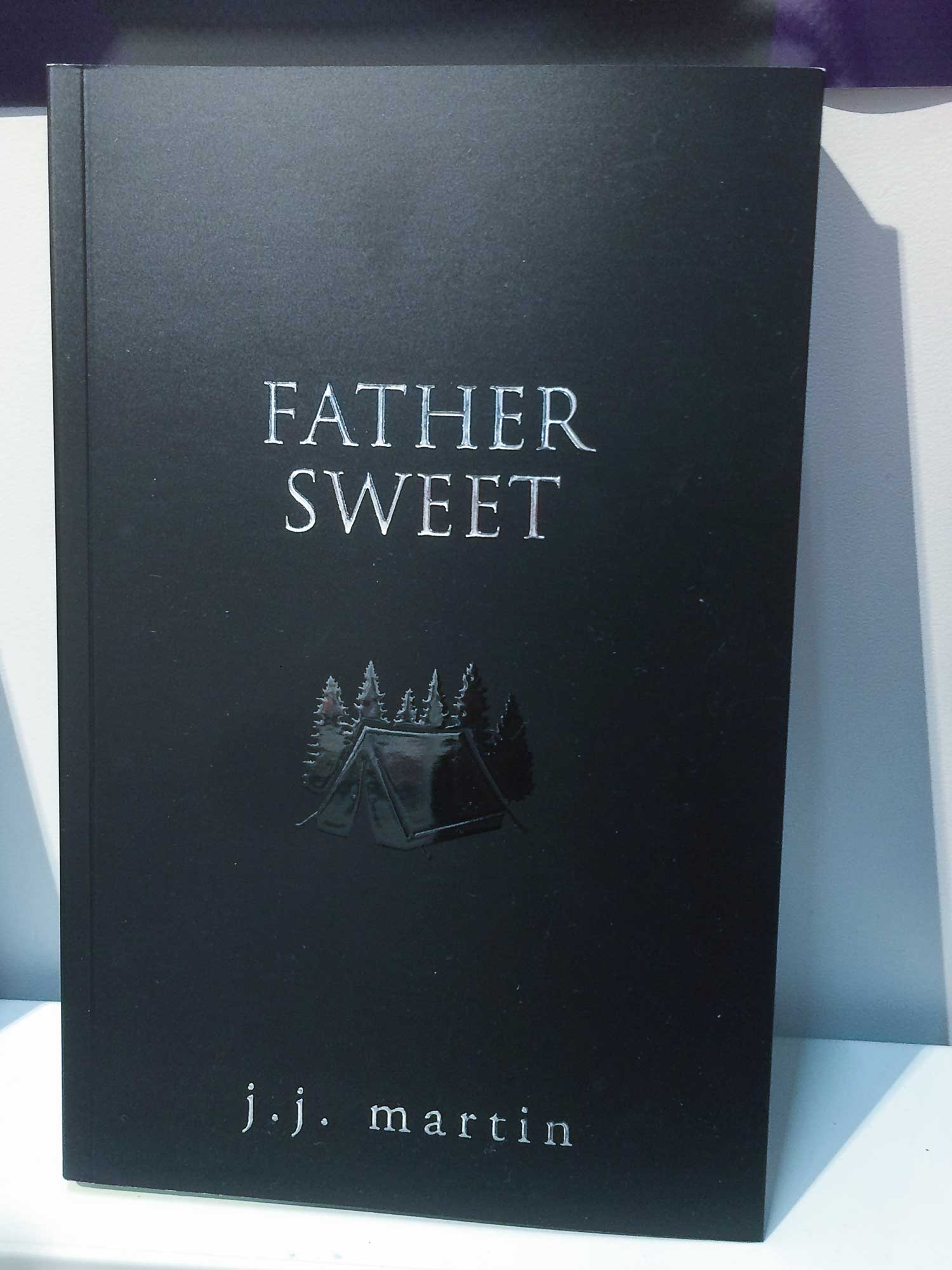 This book cover for "Father Sweet" is black with silver typography and a gloss raised print of a simple tent in the woods. It appears in Dundurn PR Ltd