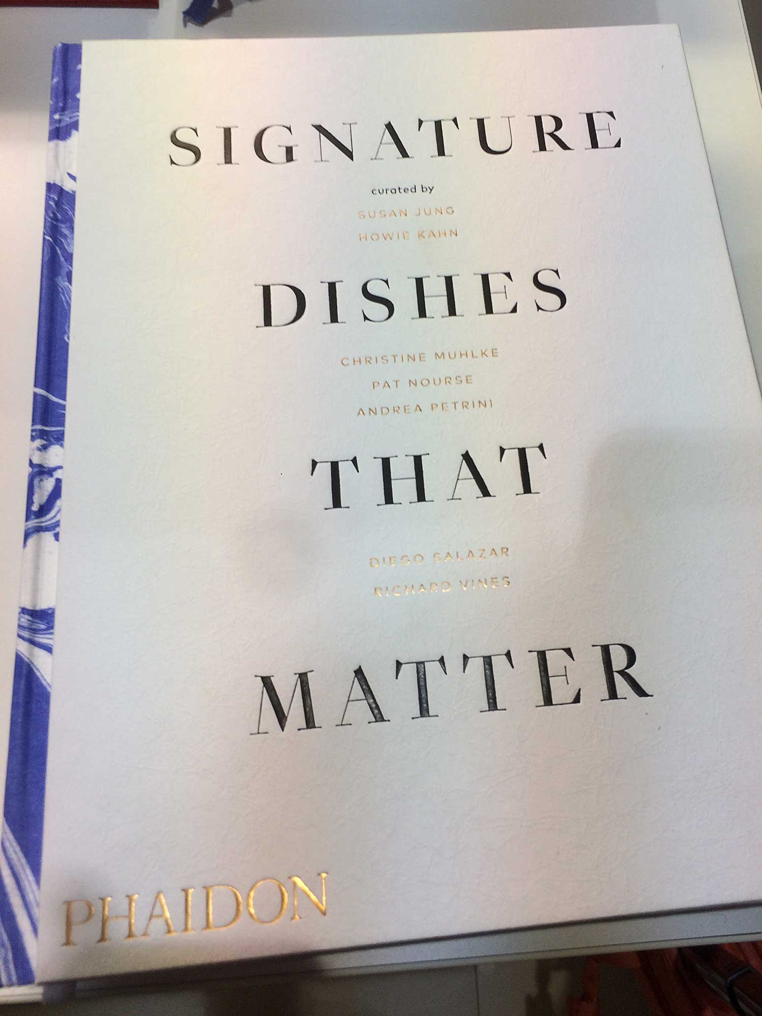 signature-dishes-that-matter-cover Dieses Foto zeigt das Cover vom Kochbuch "Signature Dishes that Matter", ein interessanter Fund auf der Messe. Das Buch ist weiß mit schwarz und gold Beschriftung. Der Buchrücken ist mit einem blau-weißem marmorierten Papier bezogen.