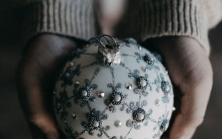 Photo © Annie Spratt on Unsplash. Photo of women's hands holding a white Christmas ornament that has been decorated with anthracite and pearl colored beads and glietter.