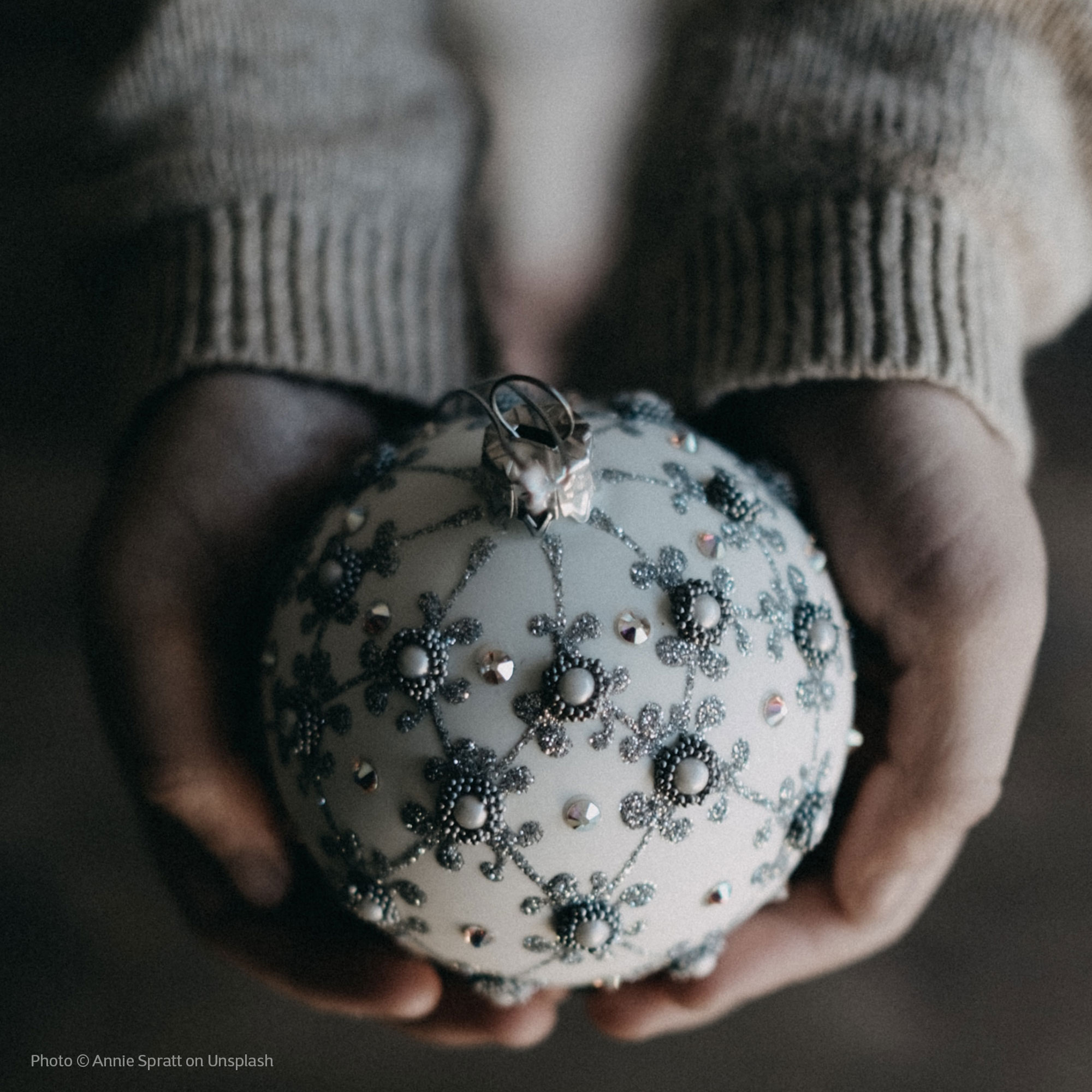 Photo © Annie Spratt on Unsplash. Photo of women's hands holding a white Christmas ornament that has been decorated with anthracite and pearl colored beads and glietter.