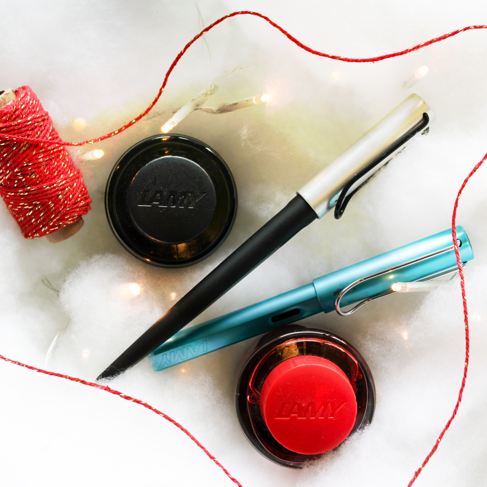 20191212 10 Presents Lamy Love Photo ©Kimberley Hoffman www.hoffman-illustrates.com Photo of 2 Lamy fountain pens, 2 ink bottles, one red, one black, red string, fluffy cloud like background with little Christmas lights
