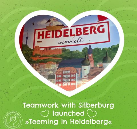 Front page of "Heidelberg wimmelt", framed with a white heart. The background is green. The caption reads: Teamwork with Silberburg launchen »Teeming in Heidelberg«.